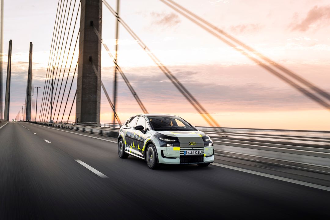 Audi A2 e-tron, prototyp w kamuflażu — przód i bok, dynamiczna jazda przez most Öresund o zachodzie słońca