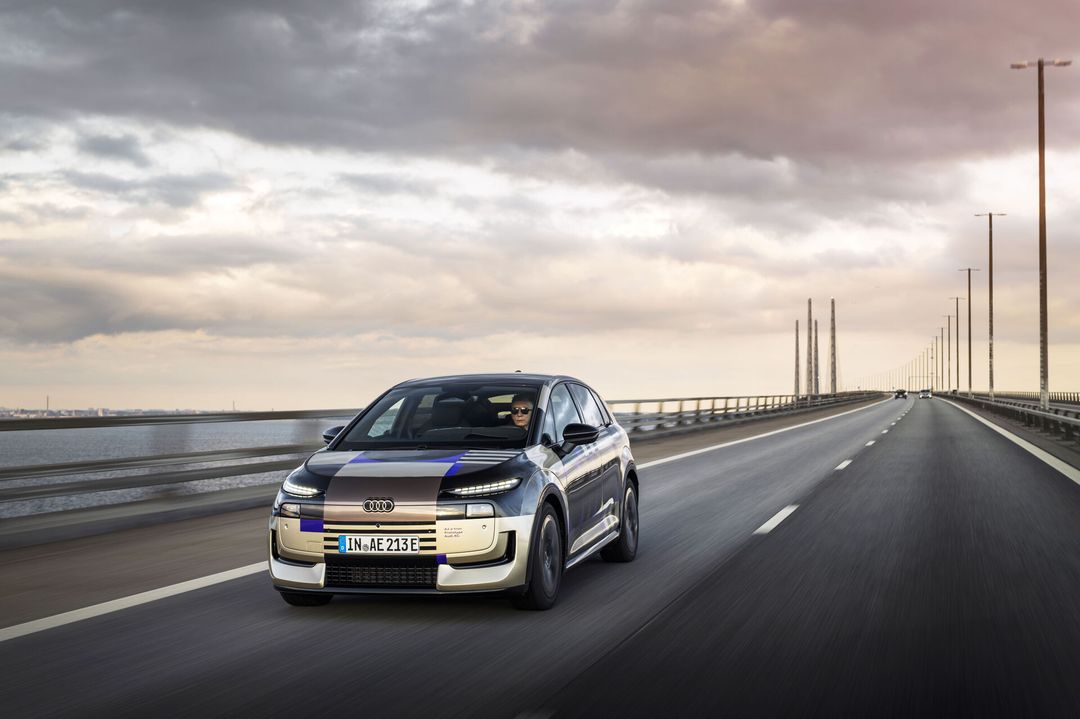 Audi A2 e-tron, prototyp w kamuflażu — przód i bok, jazda przez most pod pochmurnym niebem, kierowca po lewej stronie