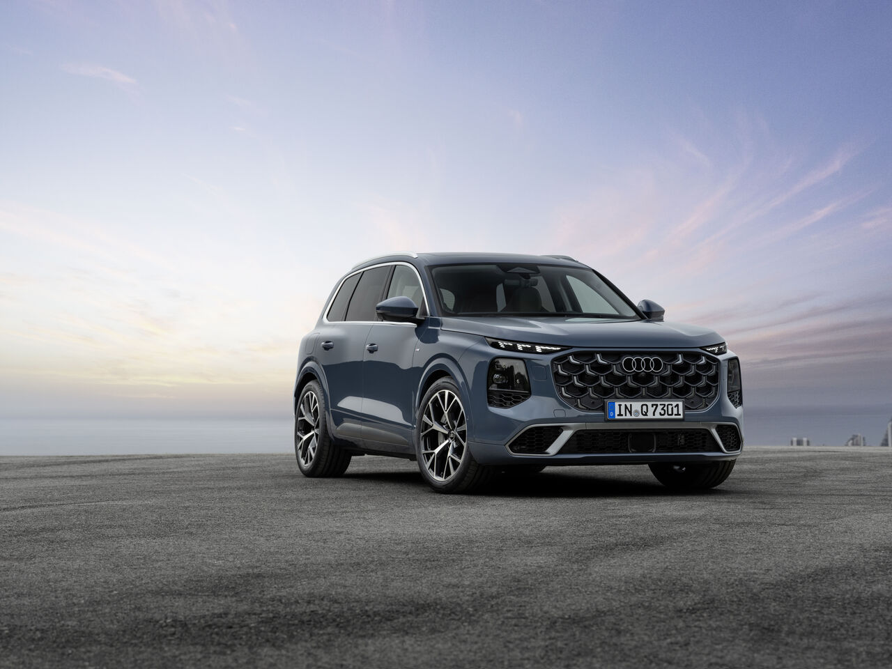 Audi Q7 III generacji — ujęcie 3/4 z przodu, kolor stalowy błękit, niemieckie tablice rejestracyjne, dramatyczne niebo w tle