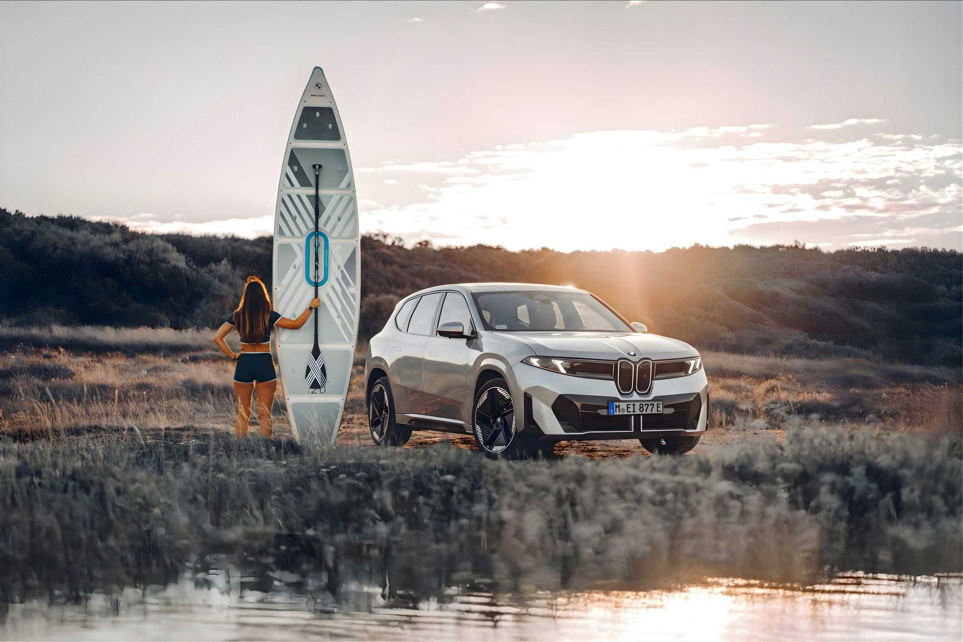 BMW X (SUV elektryczne) — przód/bok, zachód słońca nad jeziorem, obok kobieta z deską SUP