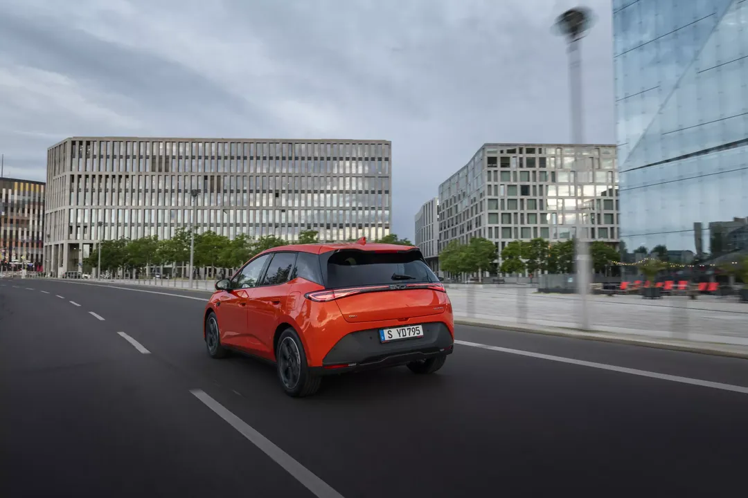 BYD Dolphin G DM-i — tył 3/4, jazda miejska, kolor pomarańczowy, rozmyte tło budynków