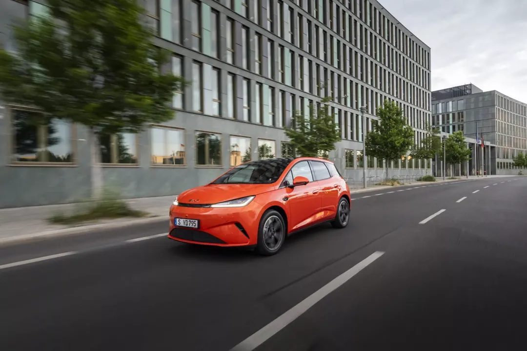 BYD Dolphin G DM-i w kolorze pomarańczowym — ujęcie 3/4 przód, dynamiczna jazda ulicą w centrum miasta