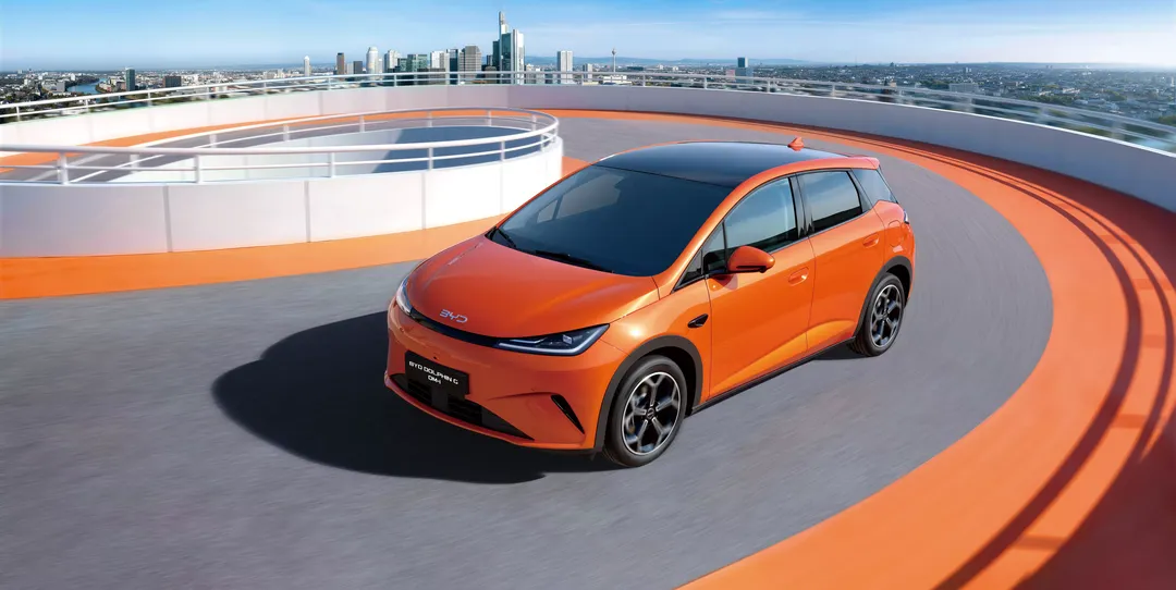 BYD Dolphin G DM-i w kolorze pomarańczowym — ujęcie 3/4 z góry na dachu spiralnego parkingu, panorama miasta w tle