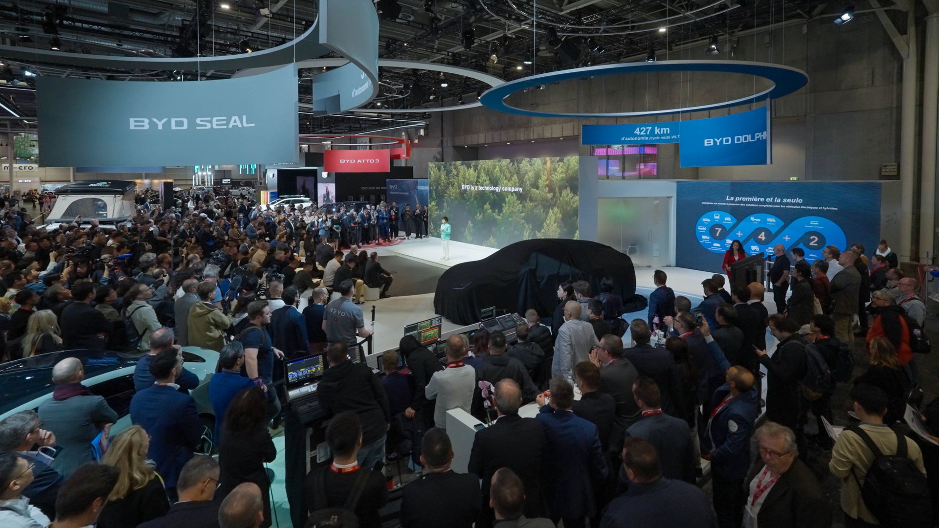 BYD — moment odsłonięcia nowego modelu pod czarną osłoną na targach motoryzacyjnych, tłum zwiedzających