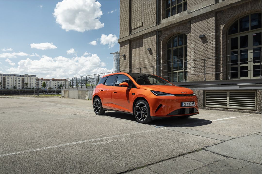 BYD — przód 3/4, pomarańczowy SUV na miejskim placu z ceglaną zabudową w tle
