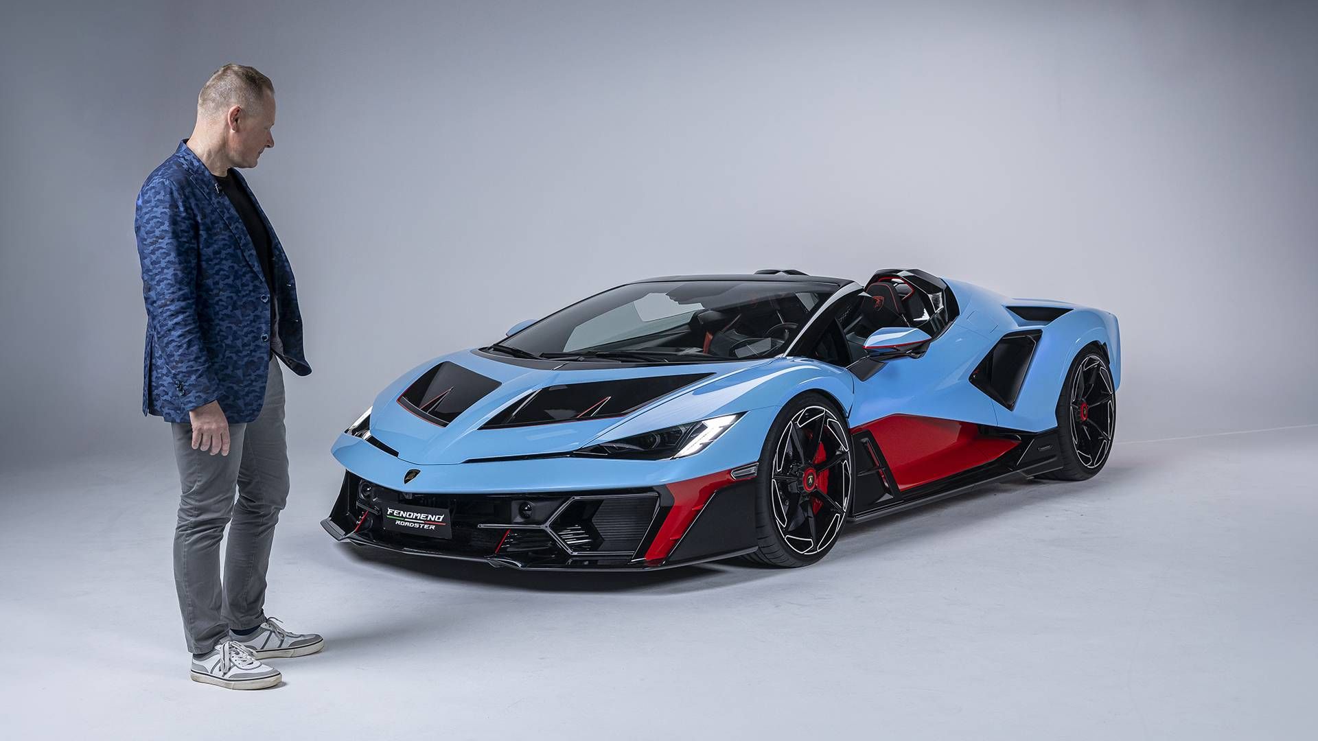 Lamborghini Fenomeno Roadster — ujęcie 3/4 z przodu, sesja studyjna, lakier błękitny z elementami z karbonu i czerwonymi akcentami, mężczyzna stojący obok dla skali