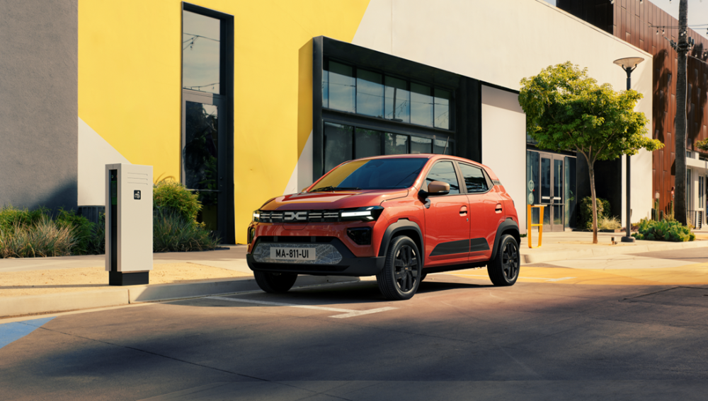 Dacia Spring — przód i bok, jazda miejska przy żółtej fasadzie budynku