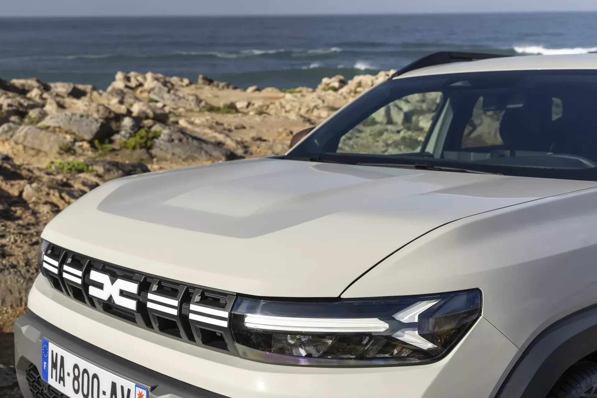 Dacia Duster — przód, zbliżenie na maskę, reflektor i grill, w tle skaliste wybrzeże i ocean
