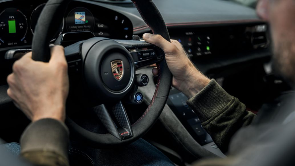 Porsche Taycan — wnętrze, kierowca trzyma kierownicę, widoczny ekran cyfrowego zegara i deska rozdzielcza