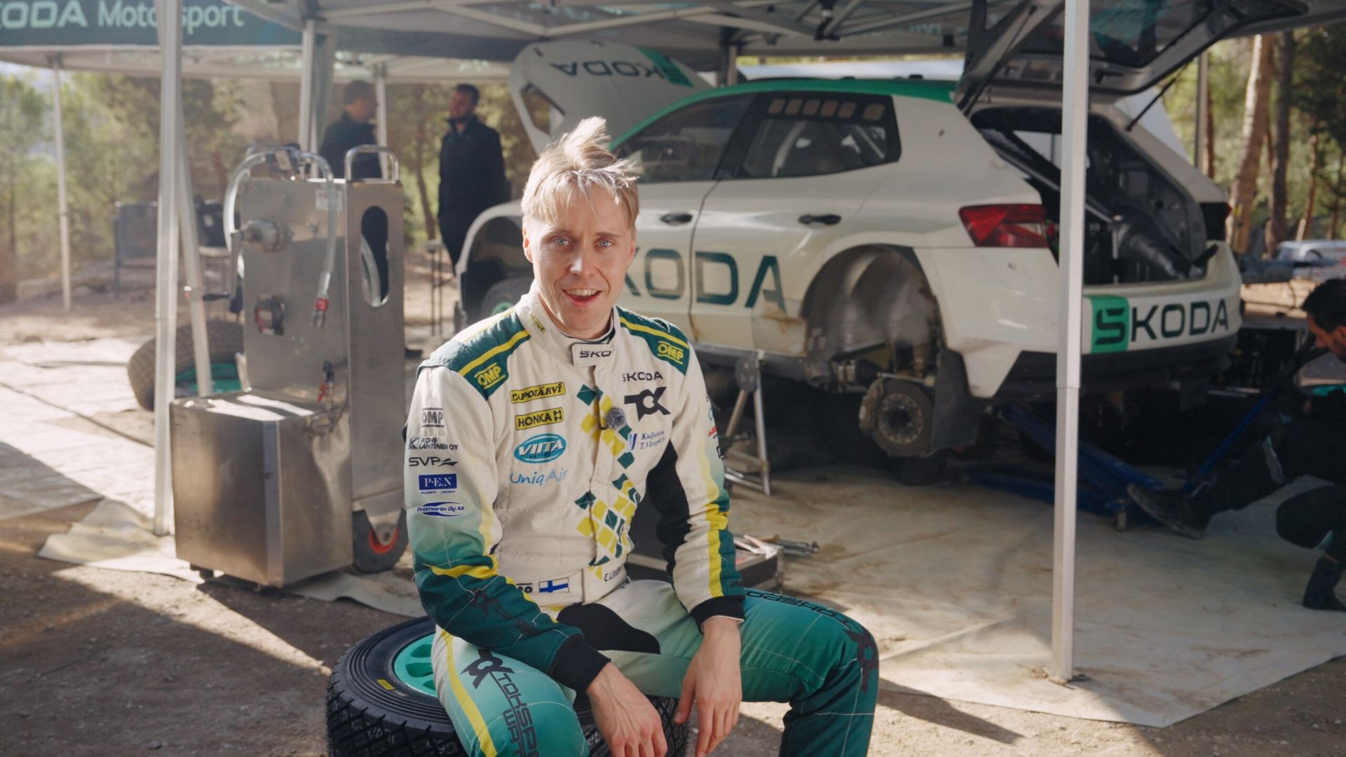 Kierowca rajdowy Škoda Motorsport w kombinezonie, w tle auto serwisowe Škoda na trasie rajdu
